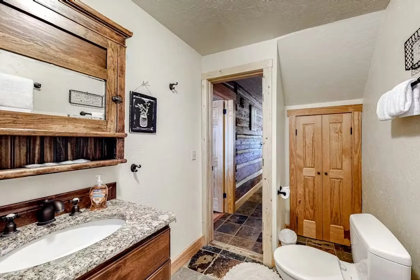 Bathroom with vanity, toilet, wooden medicine cabinet, and doorway leading to a hallway with log walls.