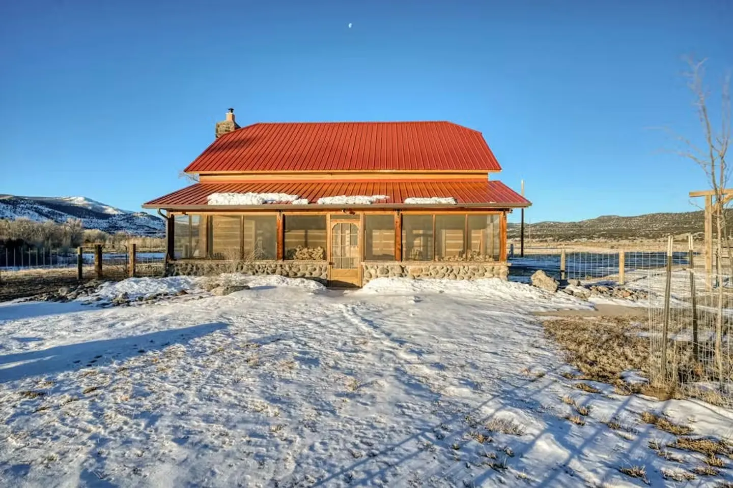 Front view of a rustic cabin with a bright red metal roof, stone foundation, and screened-in front porch, surrounded by snow-covered ground under a clear blue sky.