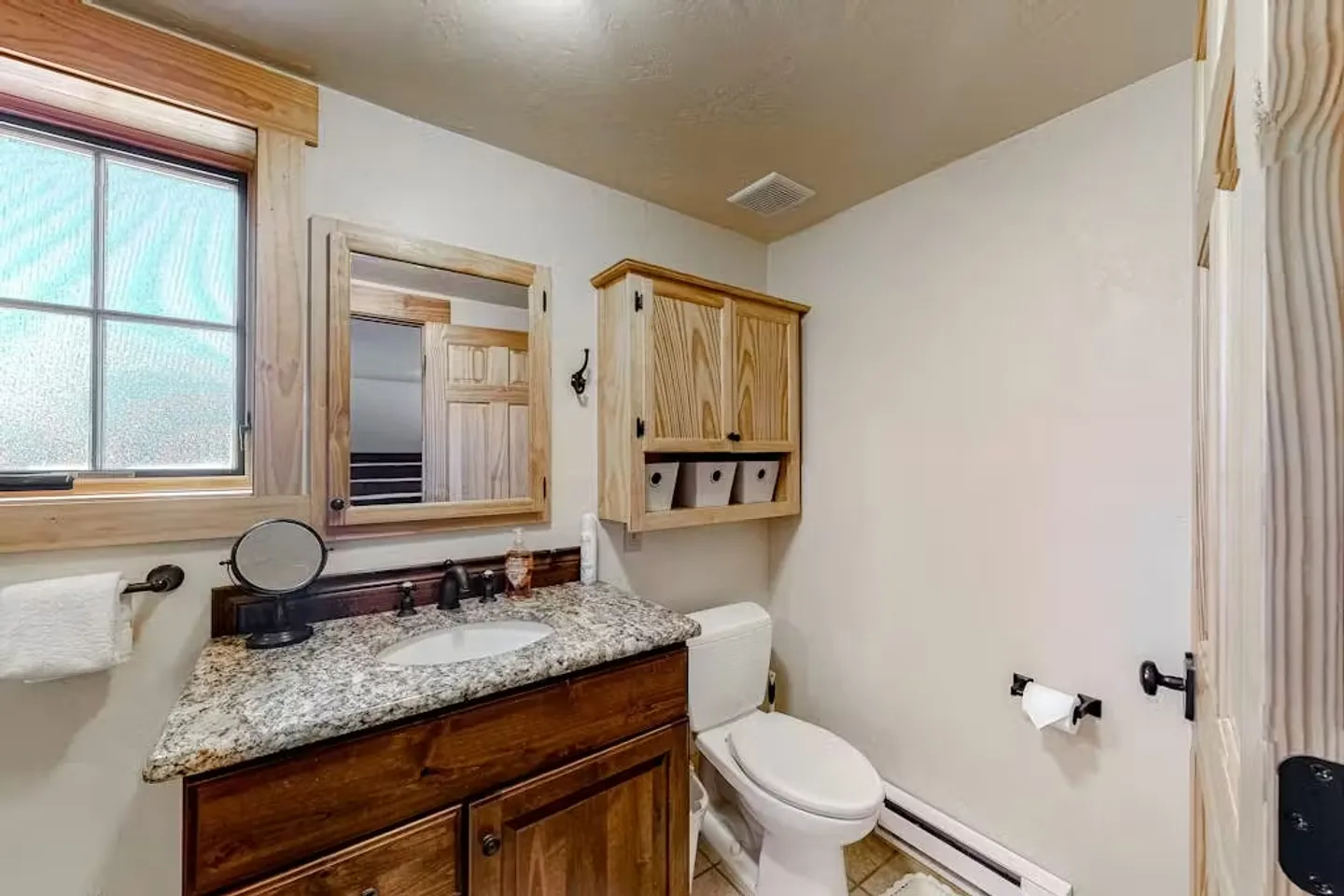 Bathroom with a wood vanity, granite-look countertop, toilet, frosted glass window, and wood wall cabinet.