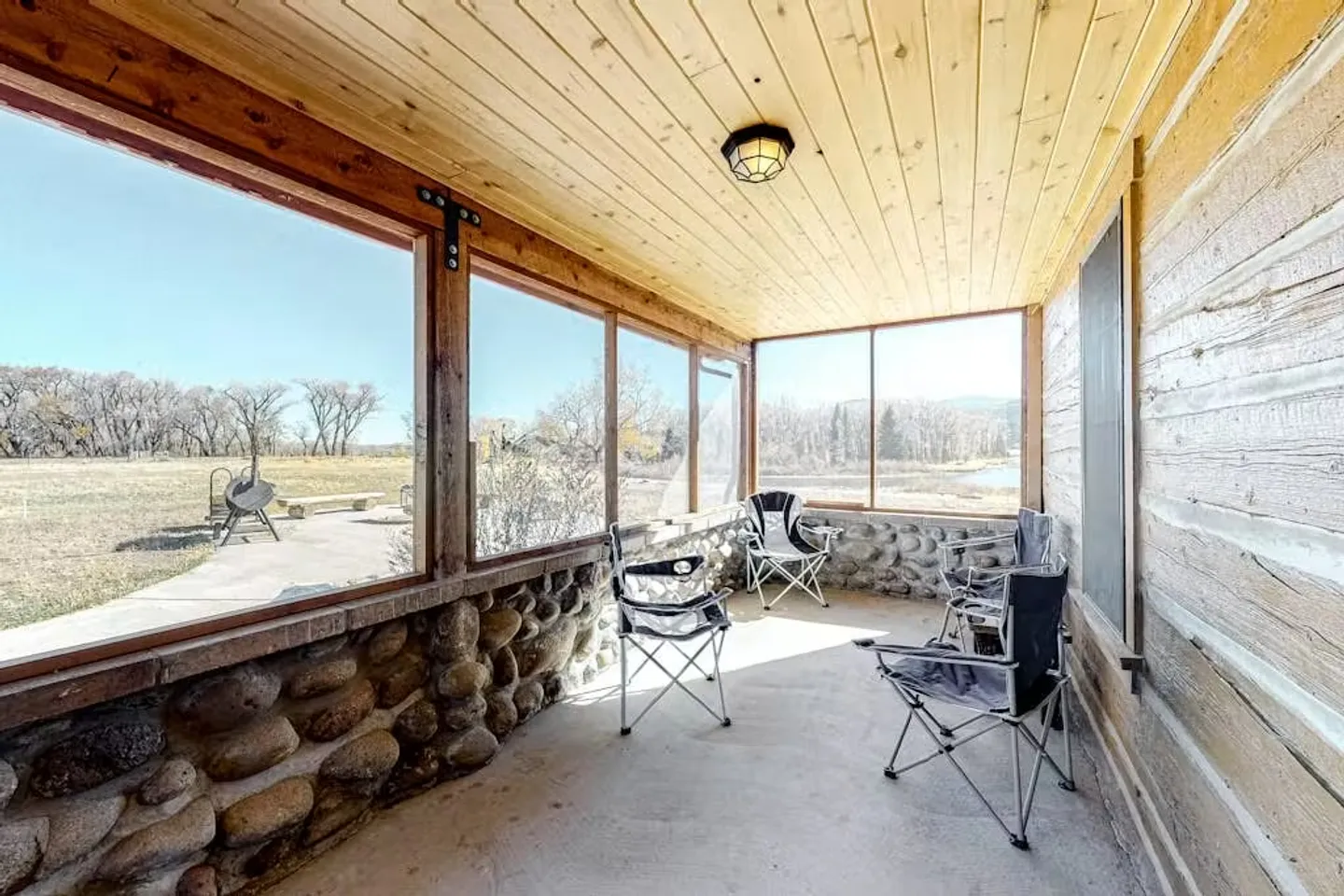 Screened porch with stone knee walls, wood paneled ceiling, and folding camp chairs looking out at an open landscape.