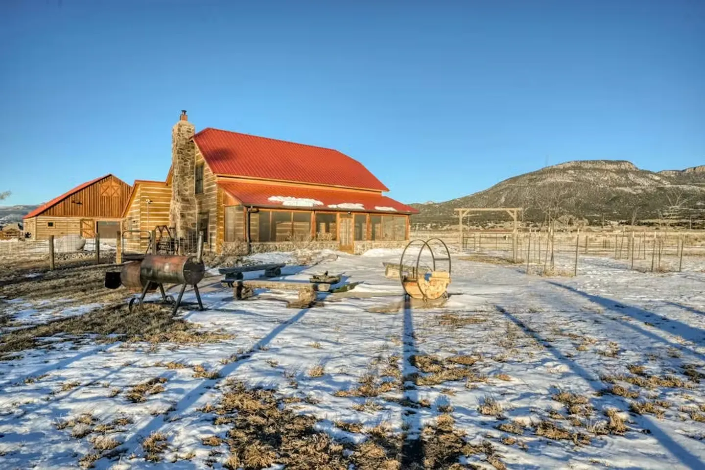 Exterior of a rustic log home with a red metal roof and outdoor seating in a snow-dusted yard against a mountain backdrop.