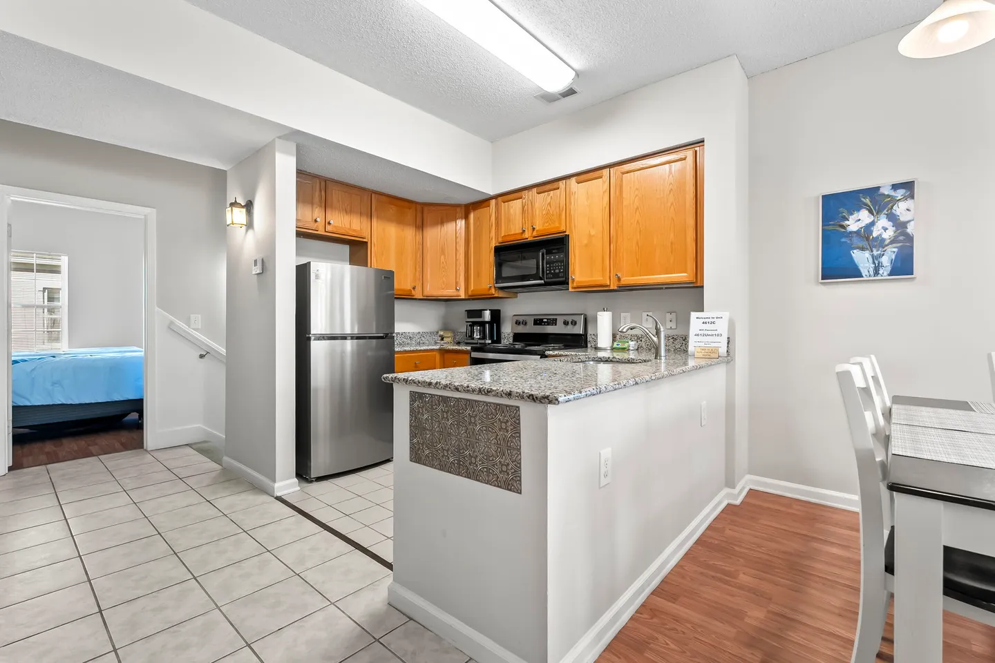 A kitchen with wooden cabinets, stainless steel refrigerator, microwave, and a breakfast bar counter adjacent to a dining area.