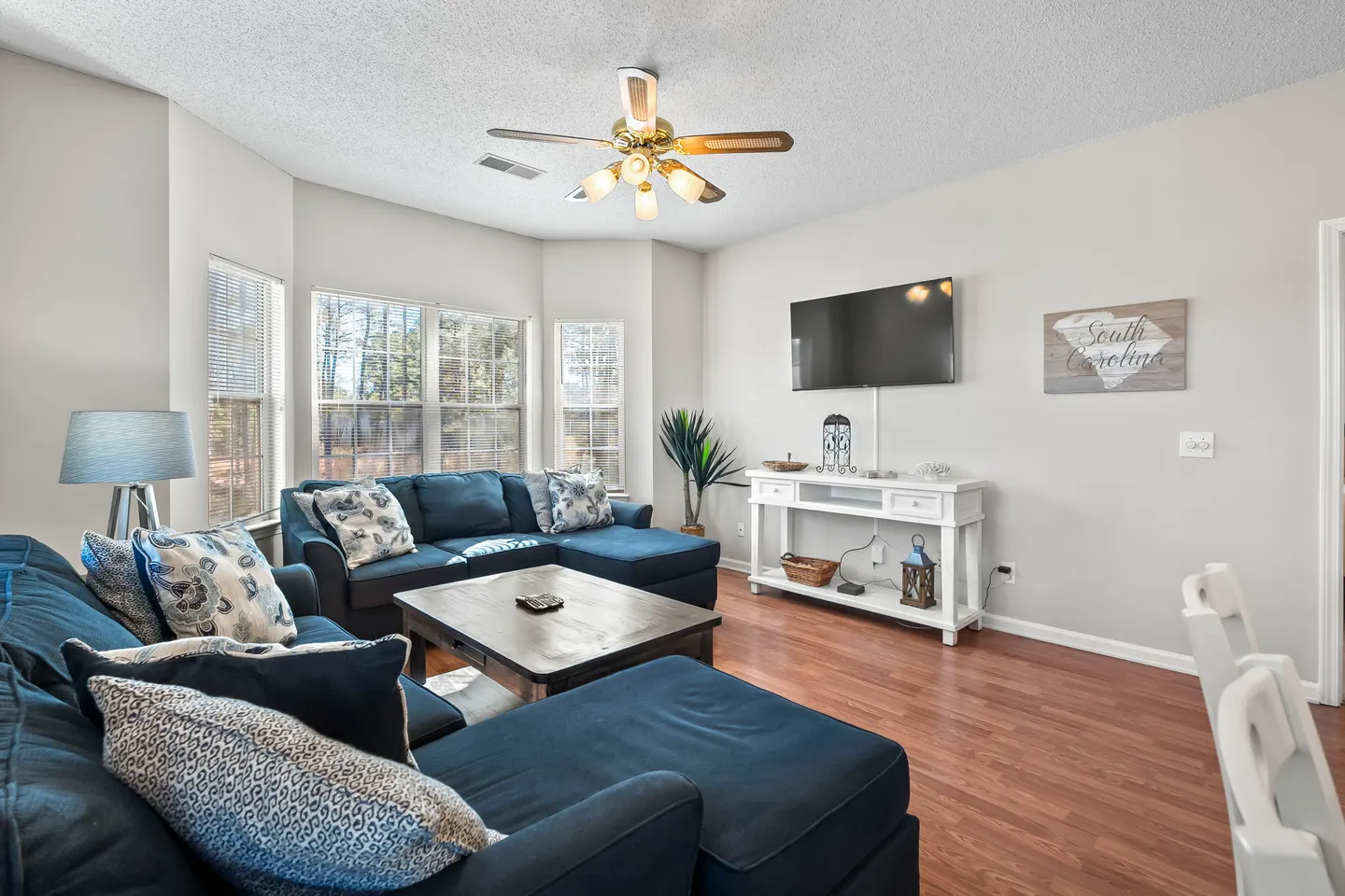 Living room with blue sectional sofas, a wood coffee table, a mounted television, and windows overlooking trees.