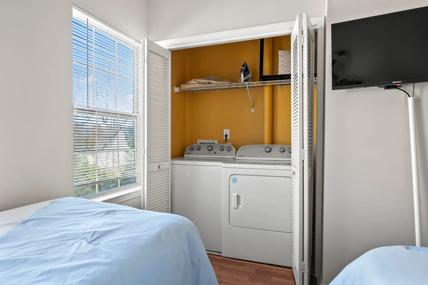 A laundry closet with white bifold louvered doors open to reveal a washer, dryer, and upper wire storage shelf located in a bedroom.