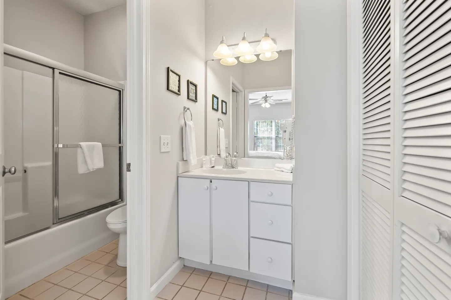 Bathroom vanity with white cabinets, a large mirror, and an adjoining room with a toilet and bathtub with glass doors.