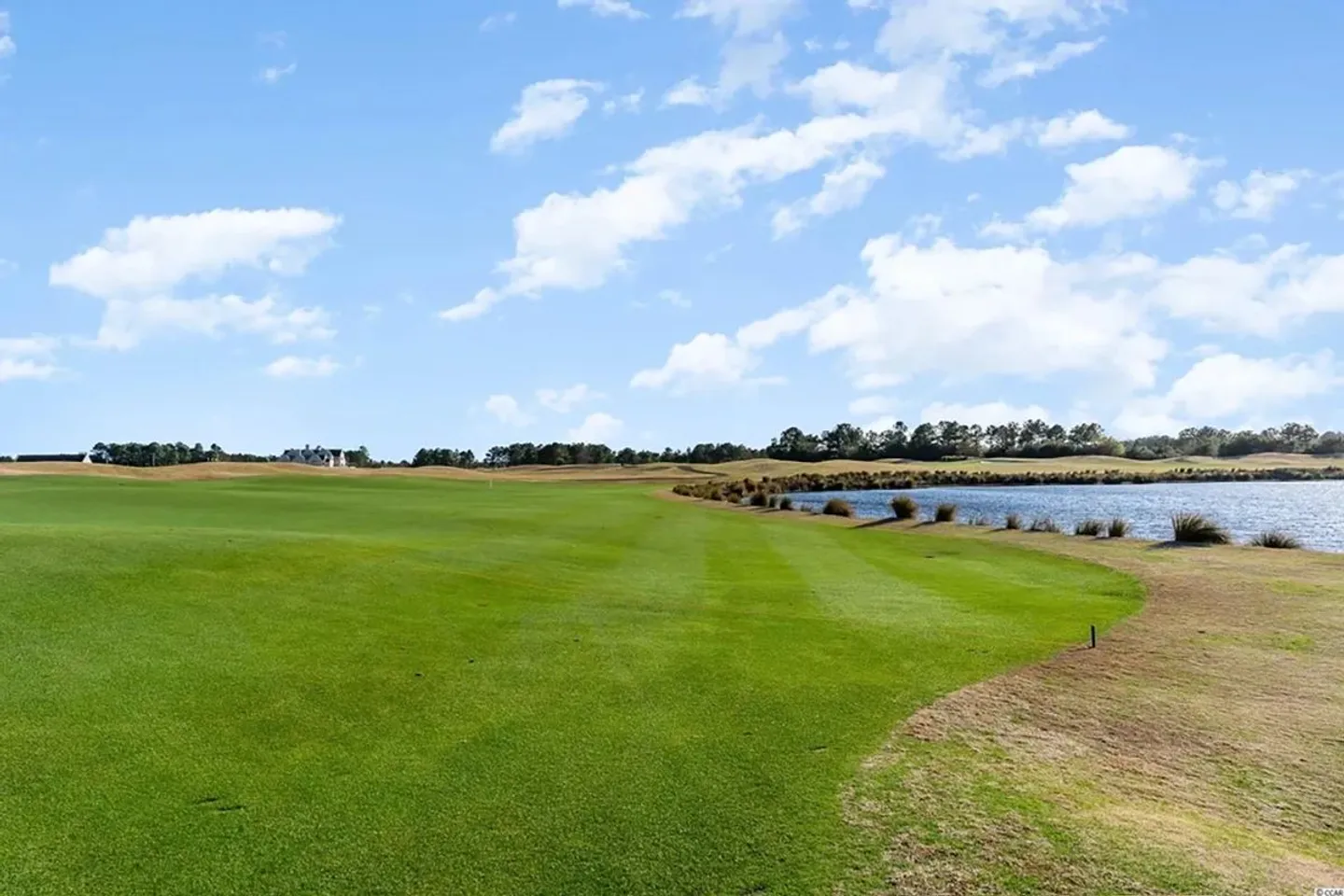 A wide view of an expansive green golf course fairway alongside a body of water under a partly cloudy sky.