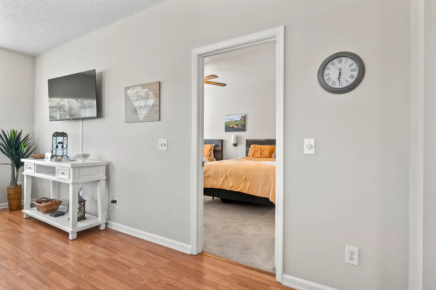 Living area with wood-look floors, a white console table, mounted TV, and an open doorway looking into a bedroom with two beds.