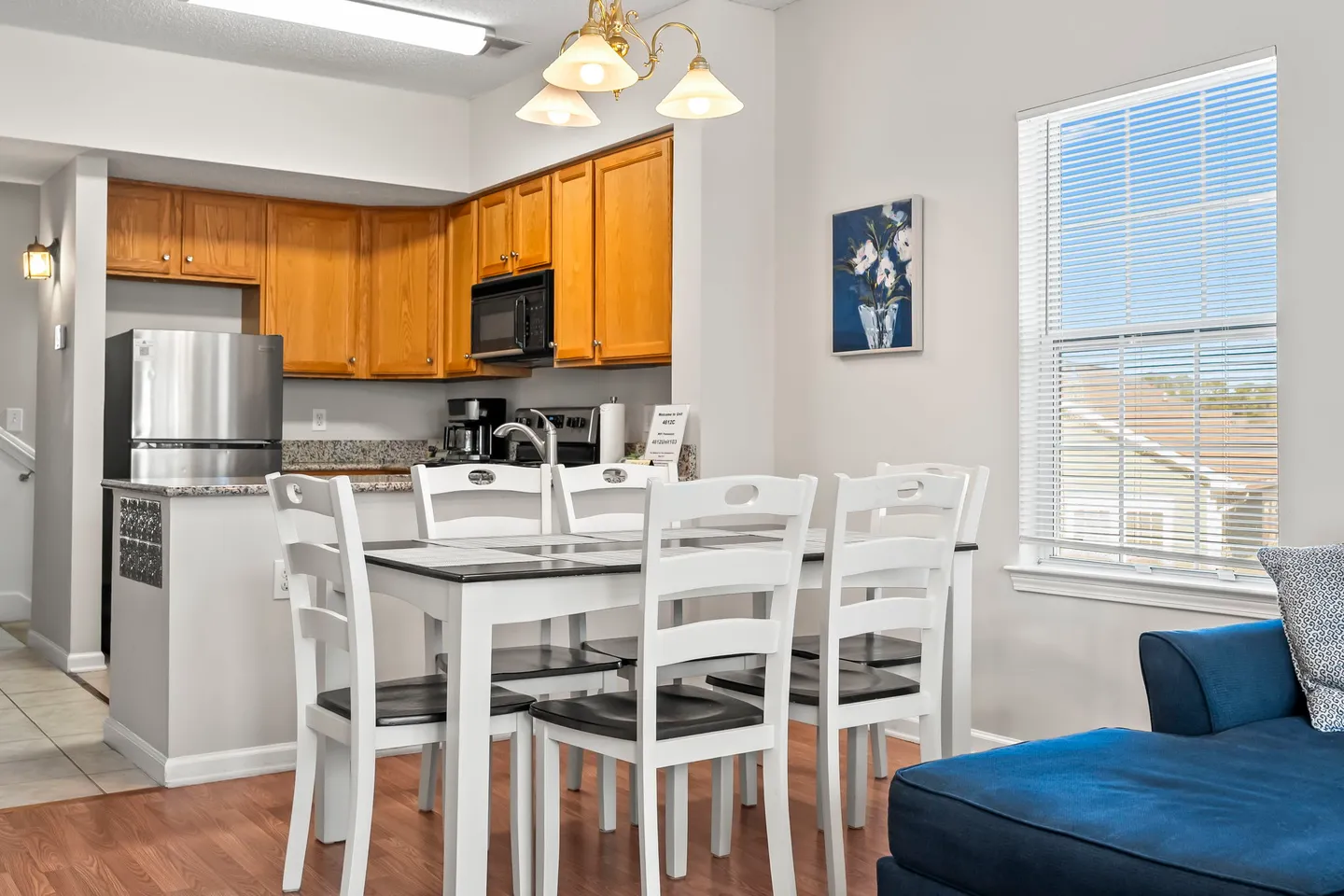 Dining area with a white wooden table and chairs adjacent to a kitchen with wood cabinetry and stainless steel appliances.