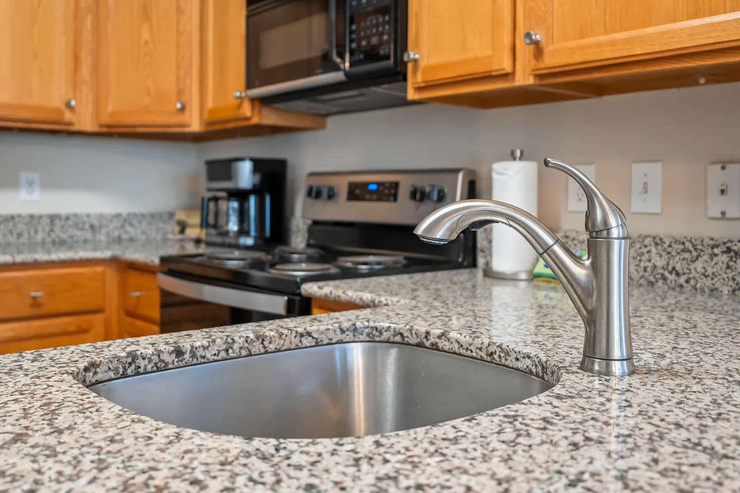 Close-up of a brushed metal gooseneck faucet and undermount sink set into a speckled granite countertop in a kitchen with wooden cabinets and stainless-steel appliances.