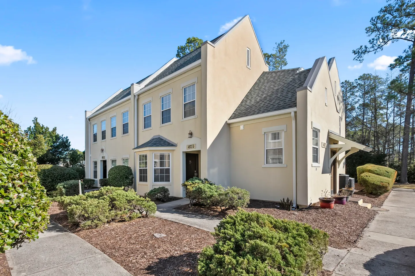 Exterior of a two-story beige townhome building under a clear blue sky, flanked by landscaped bushes and a paved walkway.