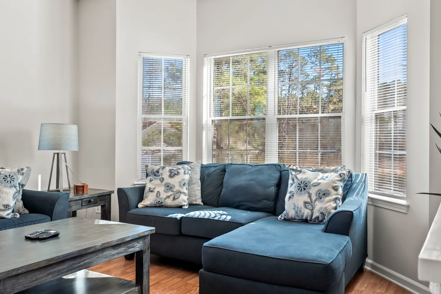 Living area featuring a blue chaise sectional sofa positioned beneath windows with white mini-blinds and a rustic coffee table in front.