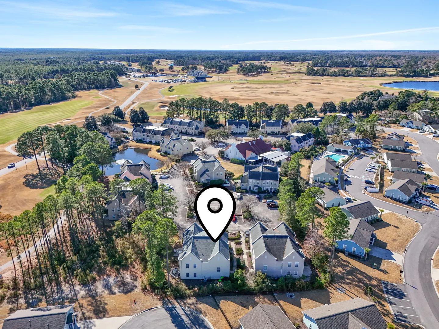 Aerial view of a residential neighborhood with townhomes and an adjoining golf course under a clear blue sky, marked with a white location pin.