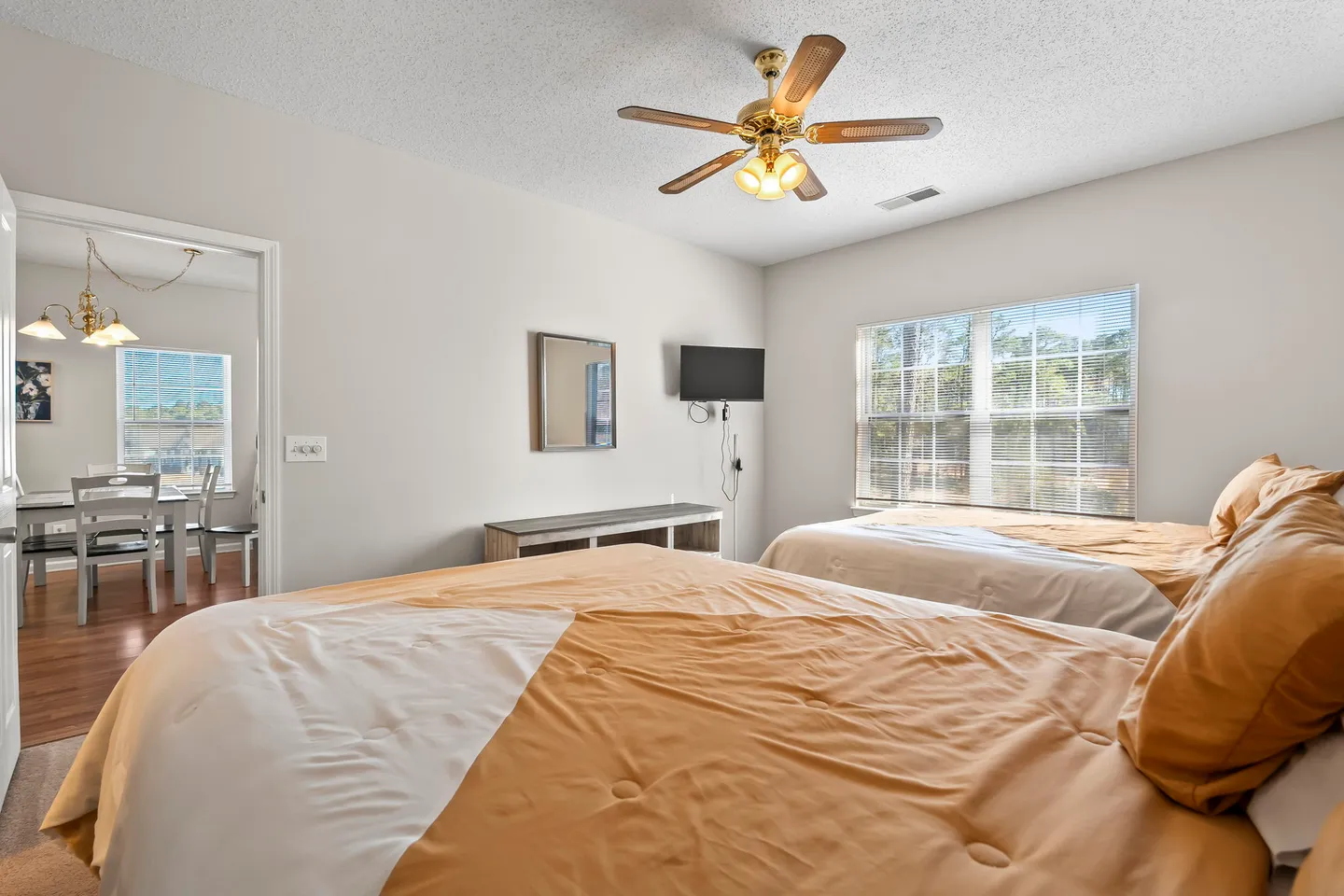 Bedroom with two beds made with tan and white bedding, a ceiling fan, a wall-mounted TV, and an open doorway leading to a dining area.