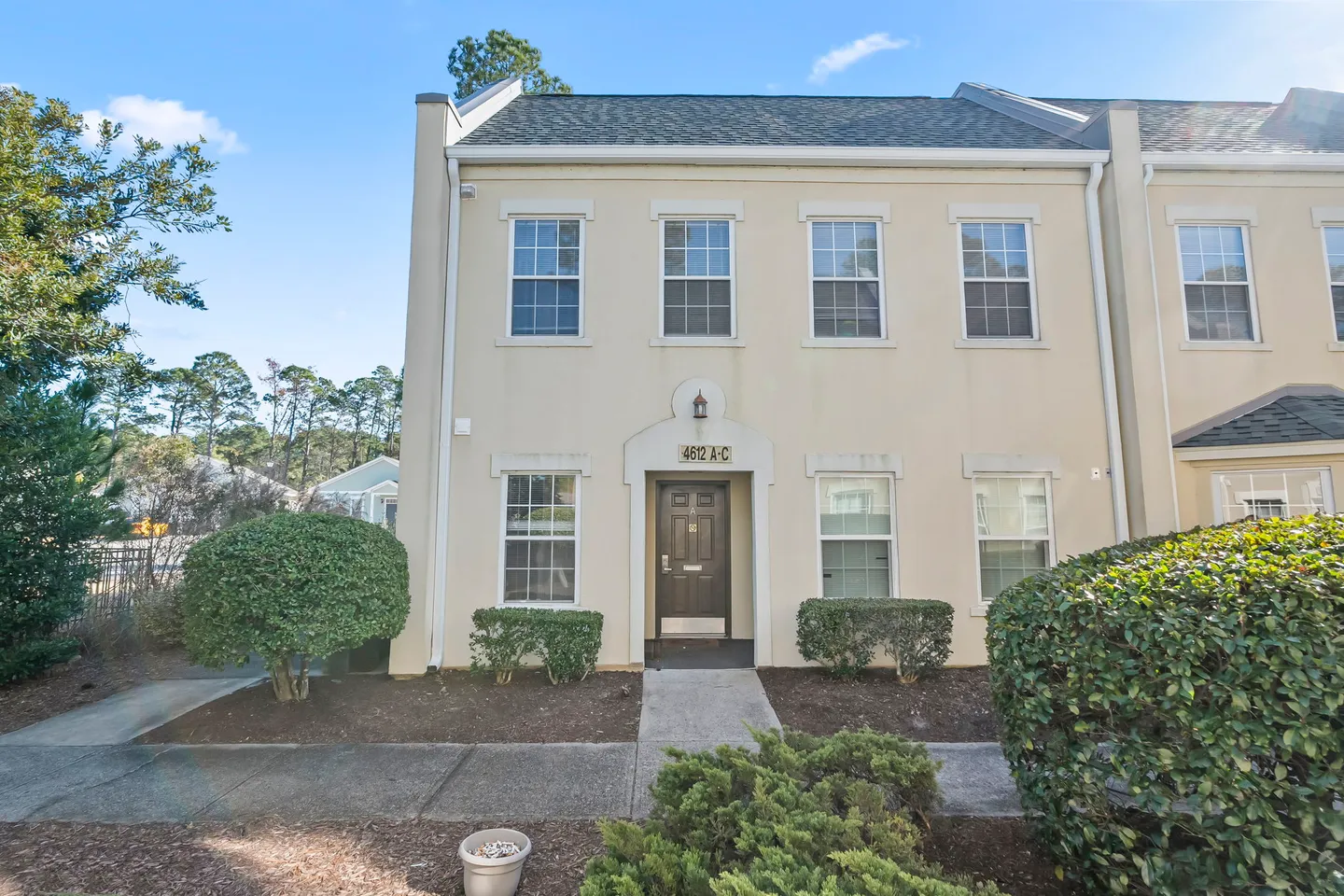 Exterior view of a two-story beige townhouse unit with a concrete walkway and landscaped bushes in front.