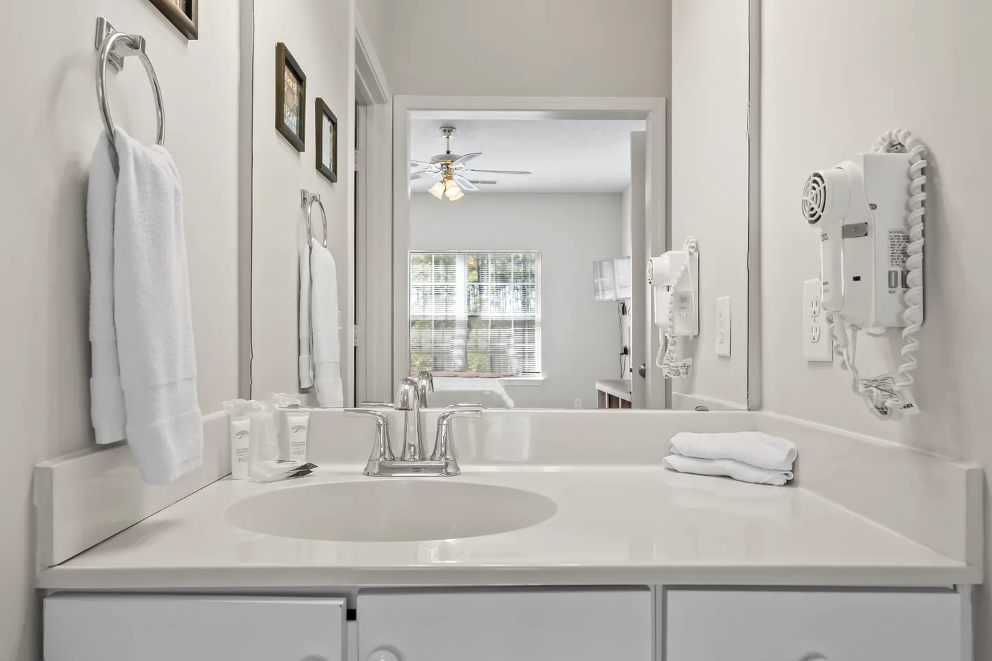 Bathroom vanity with sink, mirror, wall-mounted hairdryer, and a view into the bedroom reflected in the glass.