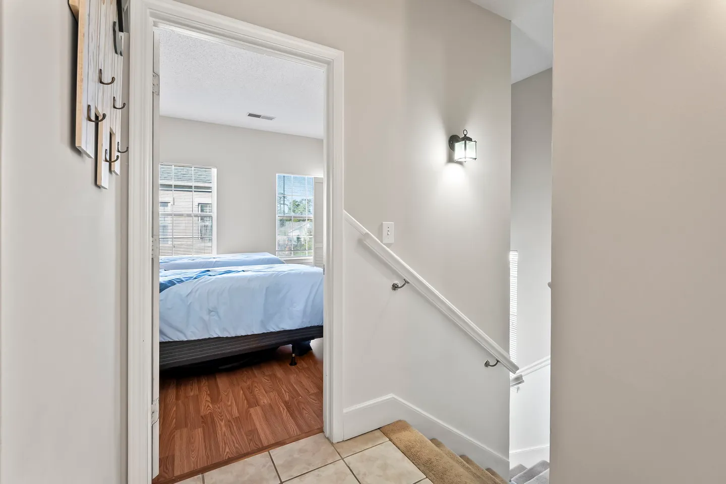 A view from a tiled landing showing an open doorway into a bedroom with wood-look floors, next to a carpeted staircase with a white handrail.