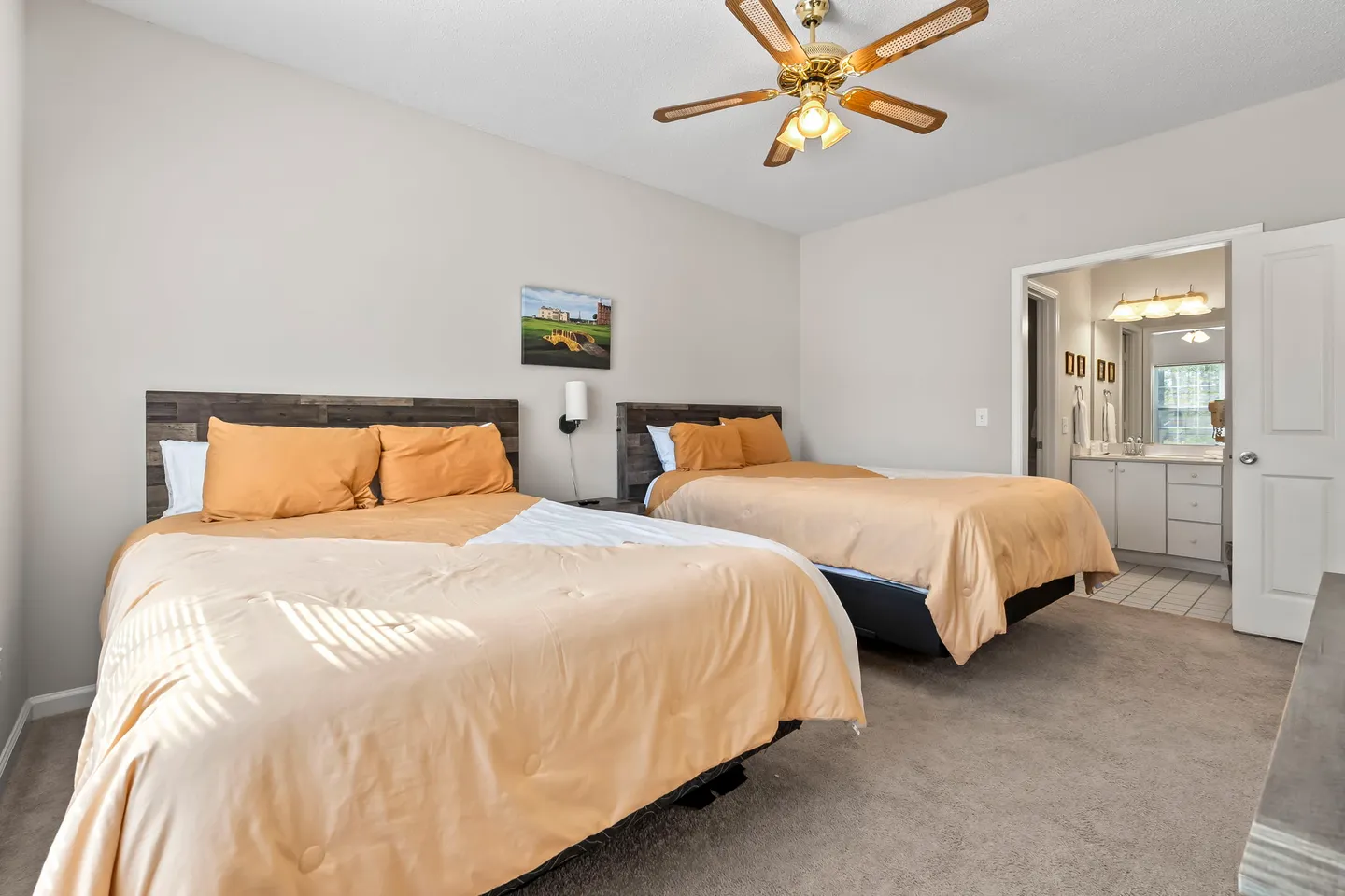 Bedroom with two beds featuring rustic wood headboards and peach bedding, carpeted floor, ceiling fan, and an open doorway to an ensuite bathroom.