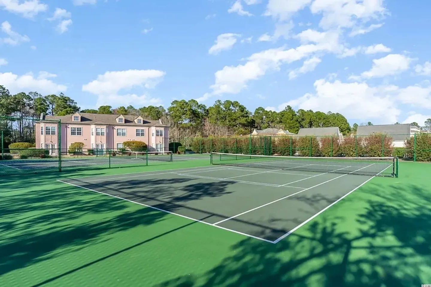Outdoor green tennis courts enclosed by chain-link fencing under a bright blue sky with residential buildings nearby.