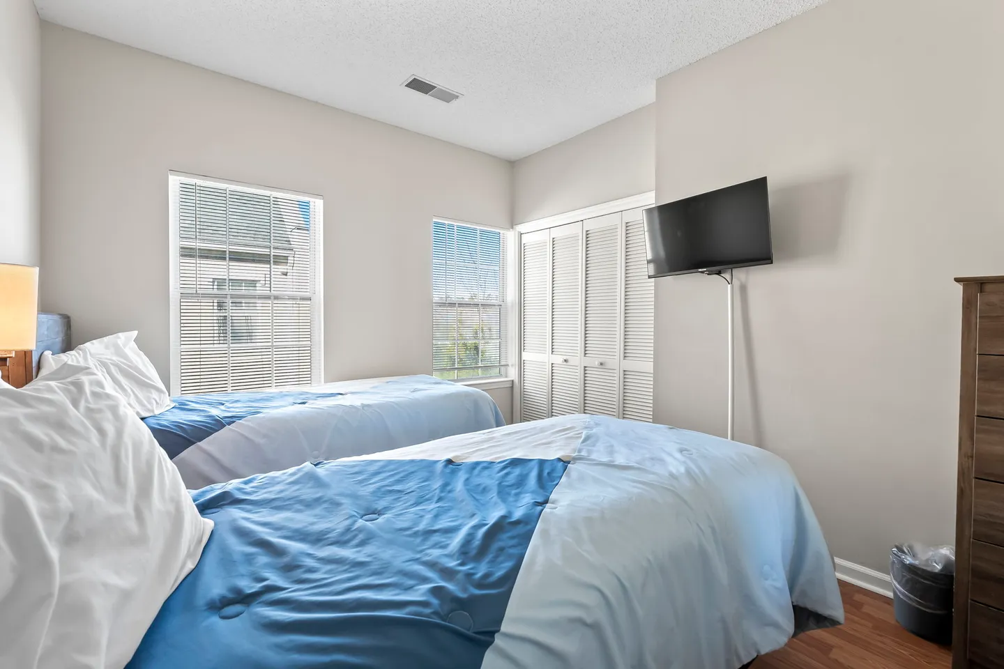Bedroom with two single beds made up with blue comforters, two windows, a louvered closet, and a wall-mounted flat-screen TV.