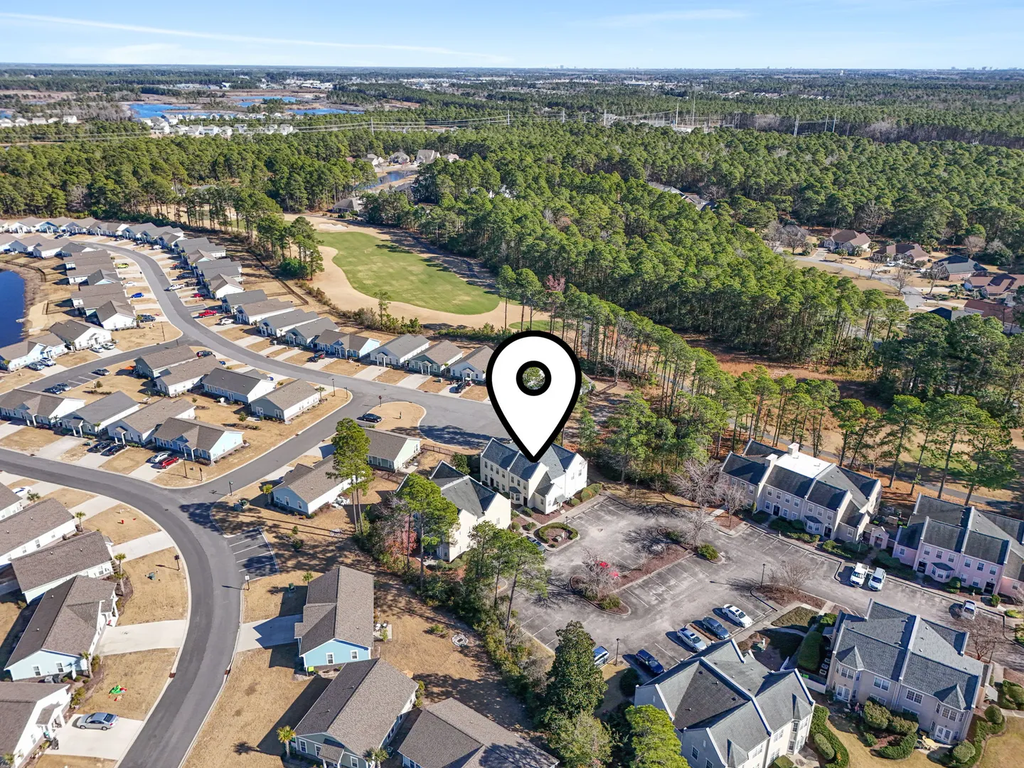 Aerial view of a residential neighborhood with townhomes, parking lots, and surrounding pine forest, marked by a location pin icon.