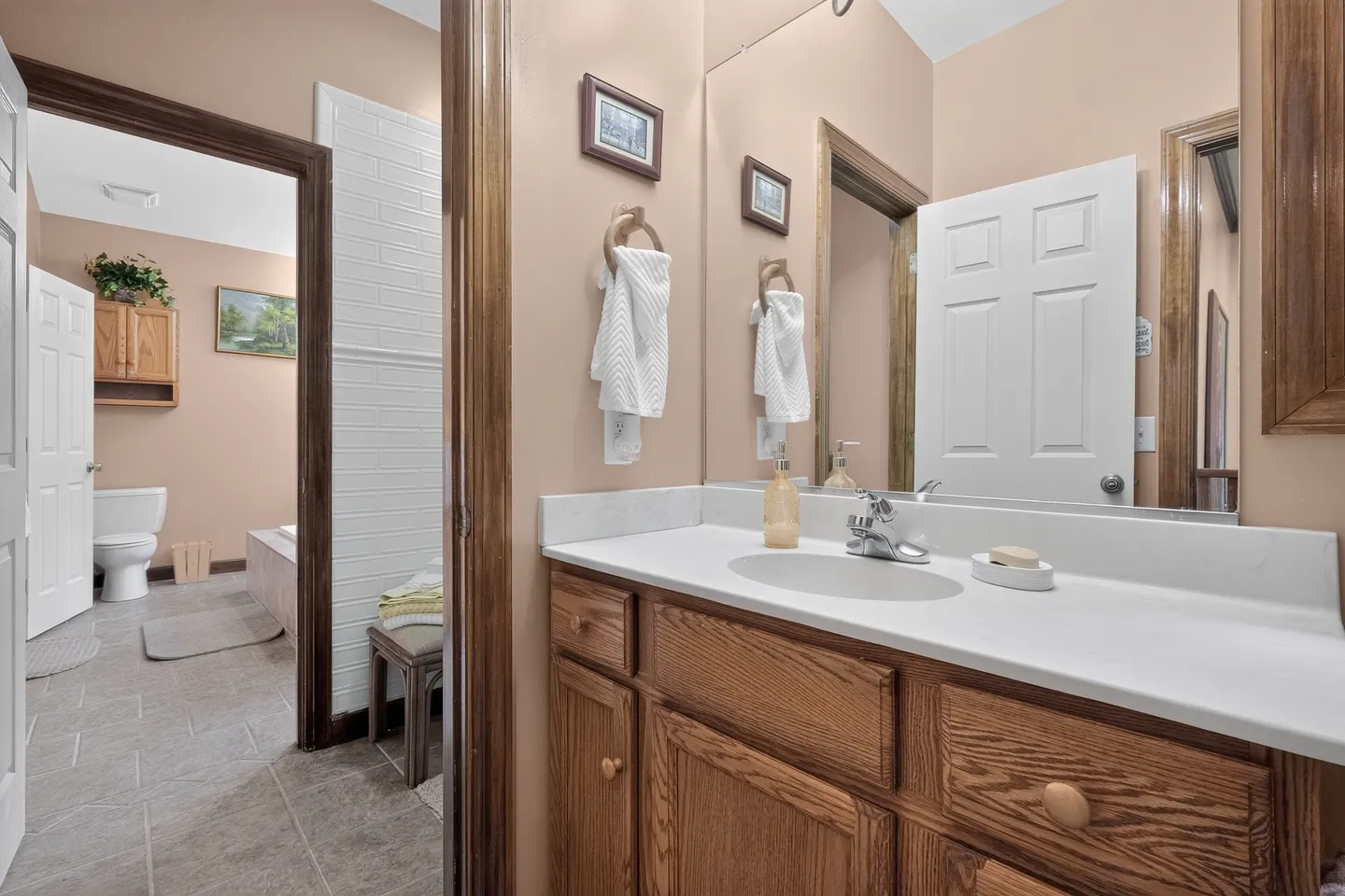 Bathroom vanity with wooden cabinets, white countertop, and mirror looking through a doorway to an inner bathroom area with tile floor, toilet, and tub.