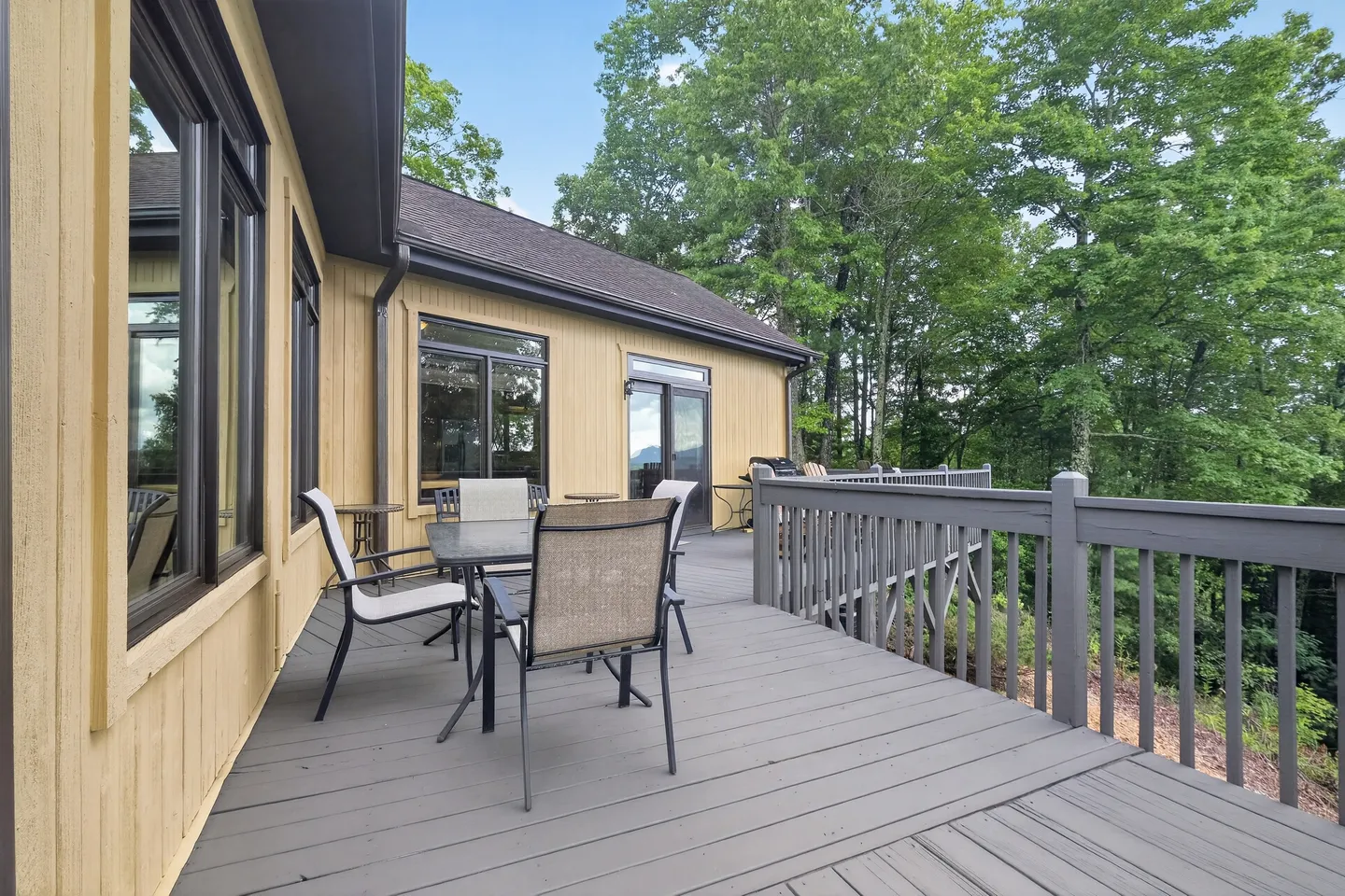 Outdoor wooden deck with outdoor dining chairs and tables next to a yellow-sided house overlooking dense green trees.