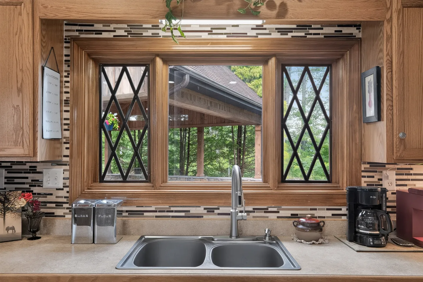 A stainless steel double kitchen sink beneath a wooden-framed window with diamond-patterned side panes looking out at an outdoor pavilion.