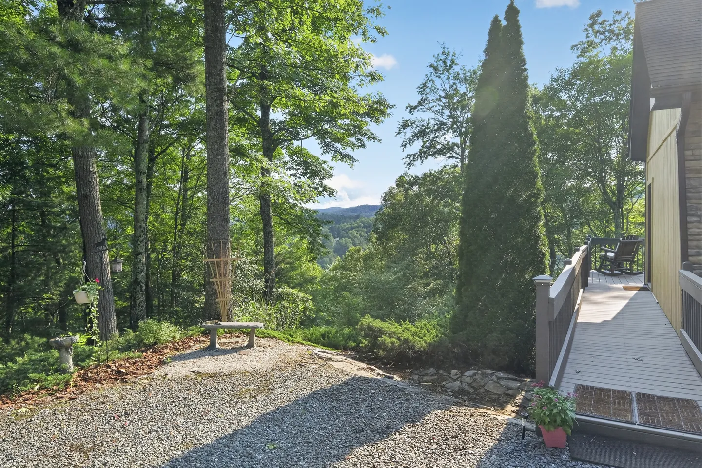 A gravel yard beside a house ramp and walkway leading to a porch, overlooking tall trees and distant mountain ridges.
