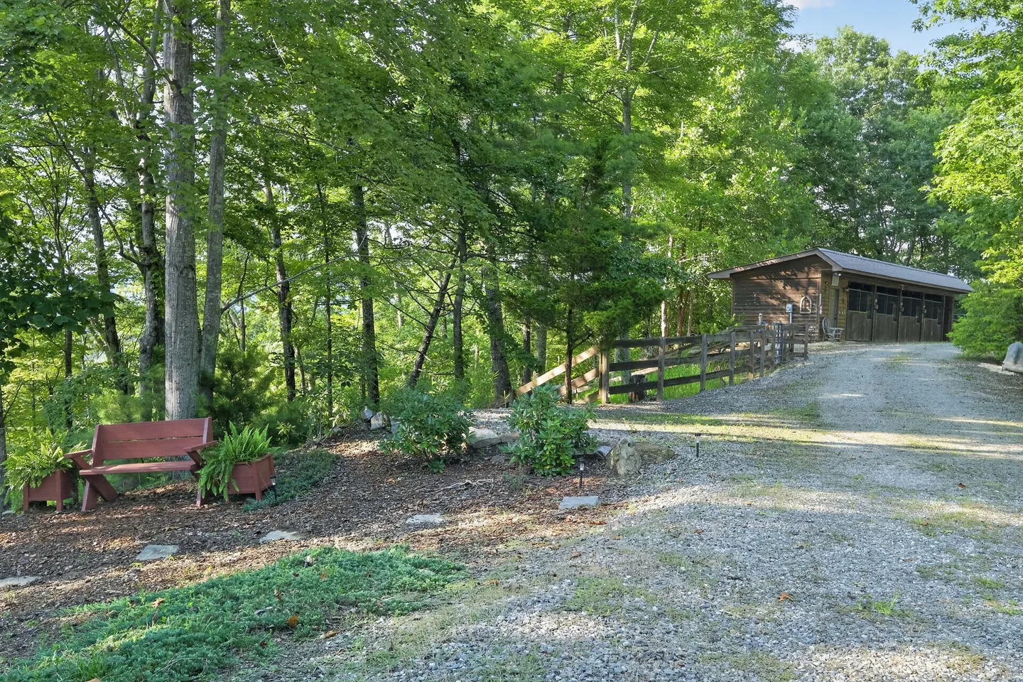 A gravel driveway leading past a bench and landscaped trees toward a wooden outbuilding or barn.