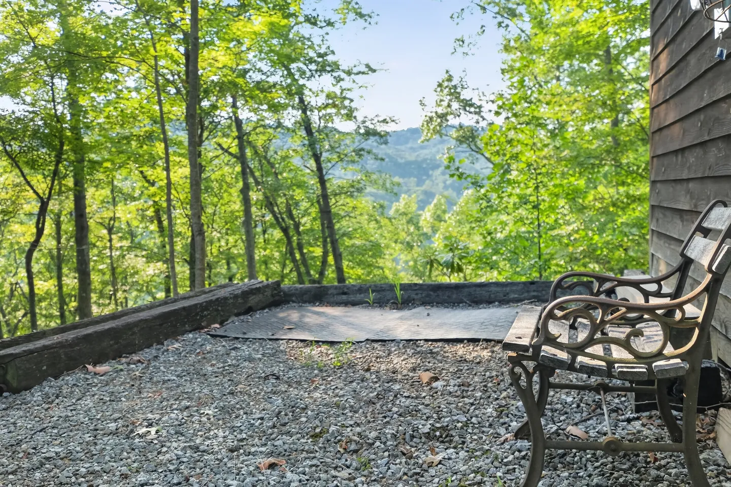 A gravel patio area with a wooden bench overlooking lush green trees and distant mountains.