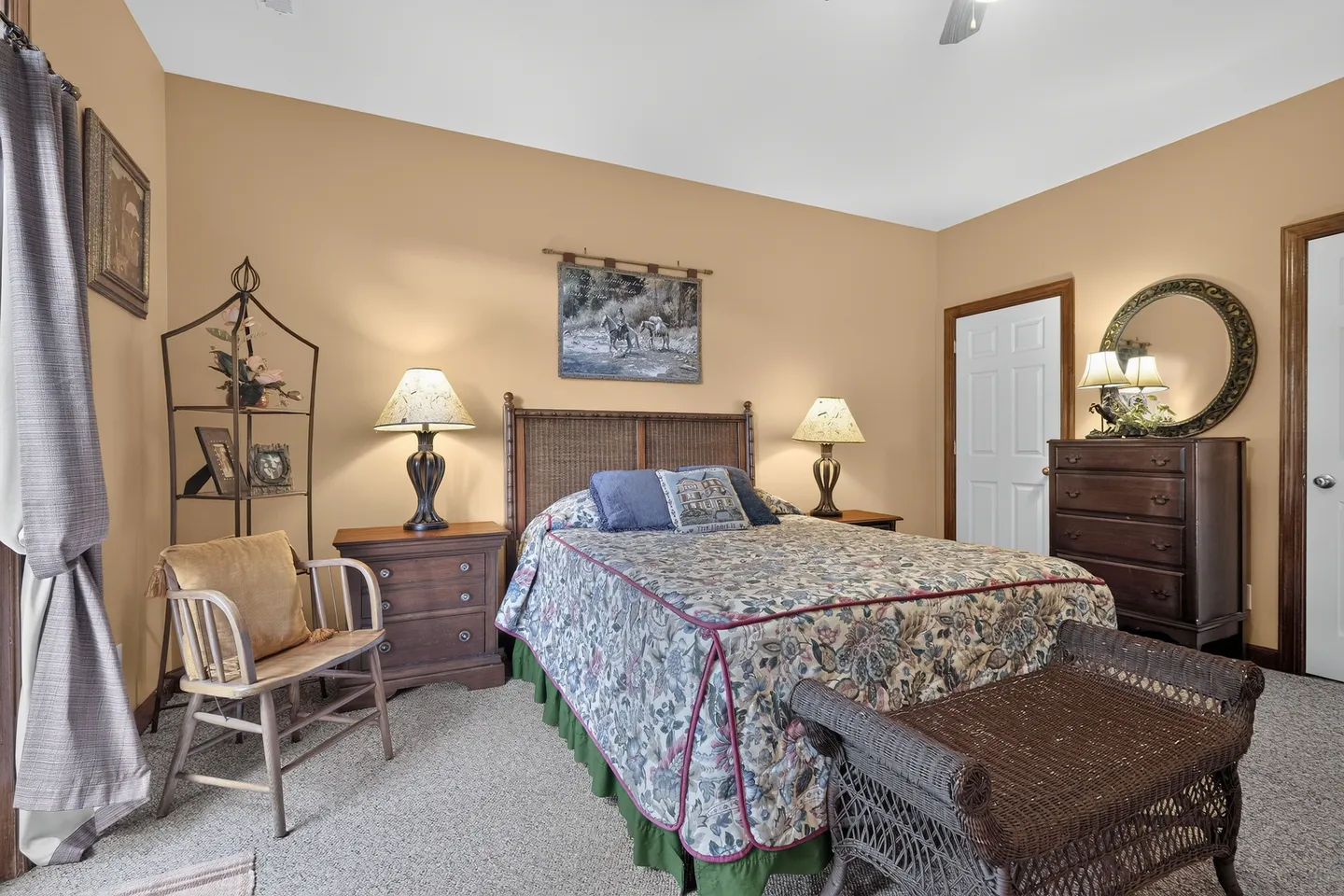 Bedroom with carpeted floor, floral bedspread on a bed with a wicker headboard, matching nightstands with lamps, a wooden dresser with a mirror, and an accent armchair.