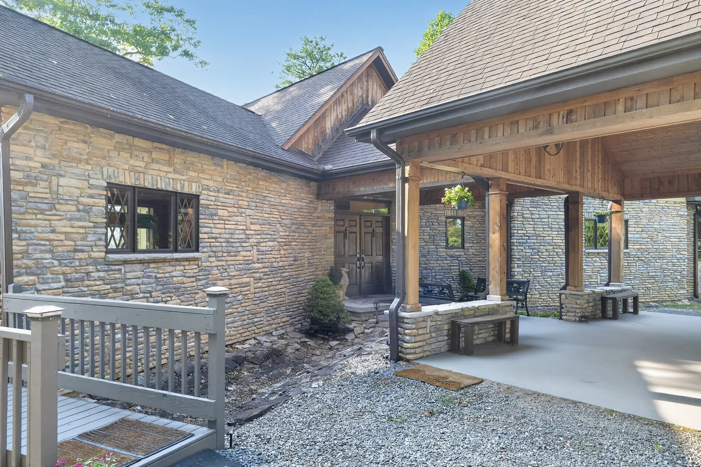 Exterior stone home entrance with a covered carport, wooden accents, and a gravel driveway.