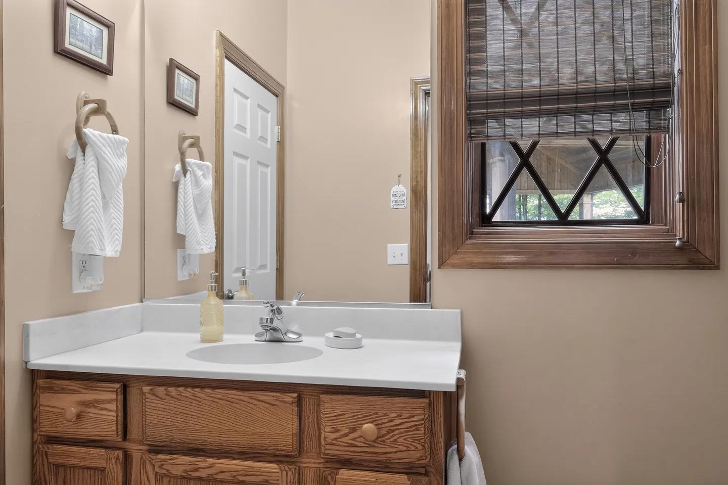 Bathroom vanity with oak cabinetry, white countertop, large mirror, and an adjacent window with cross-bracing and a woven Roman shade.
