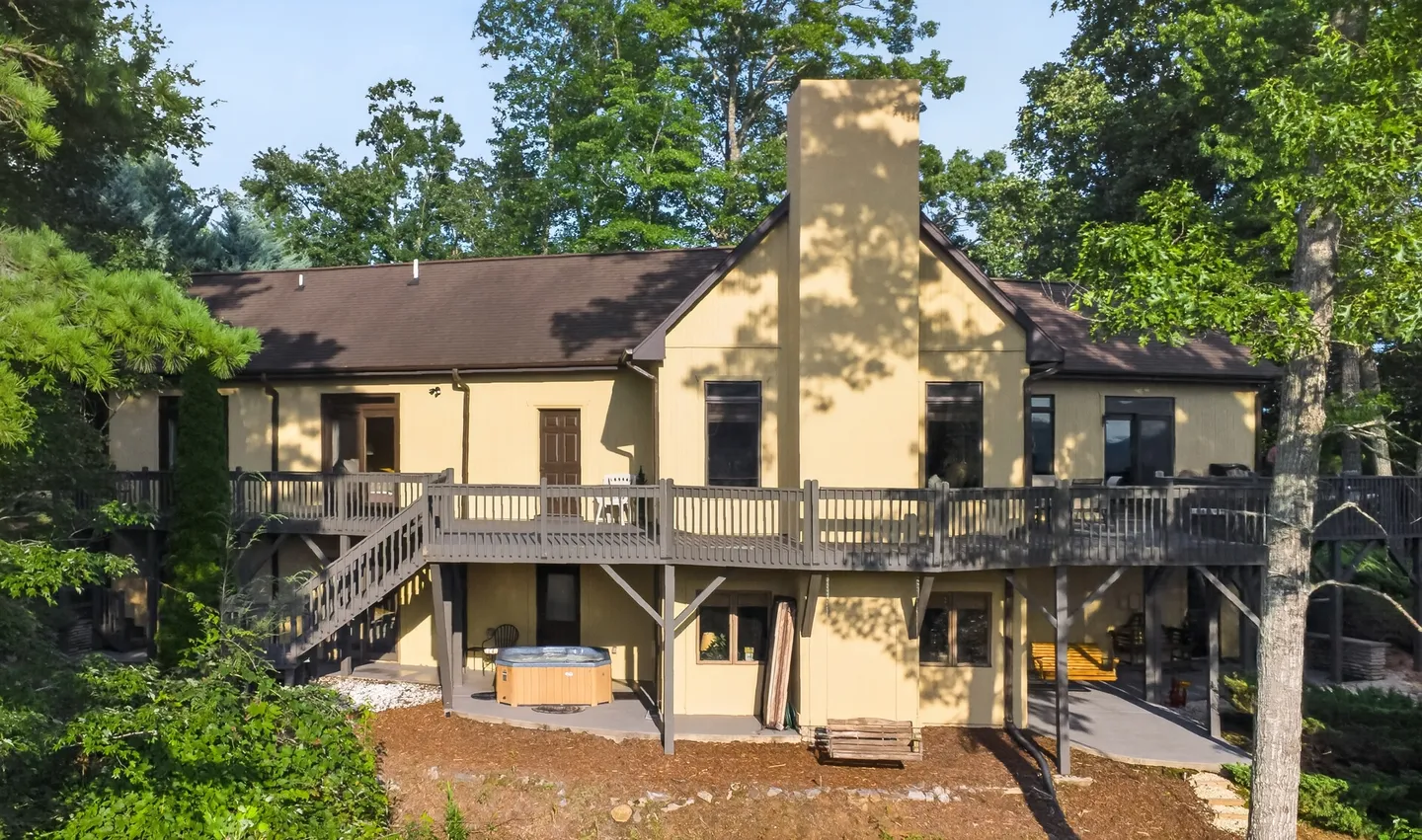 Exterior view of the back of a two-story beige house with a large wooden deck and a hot tub on the lower patio.