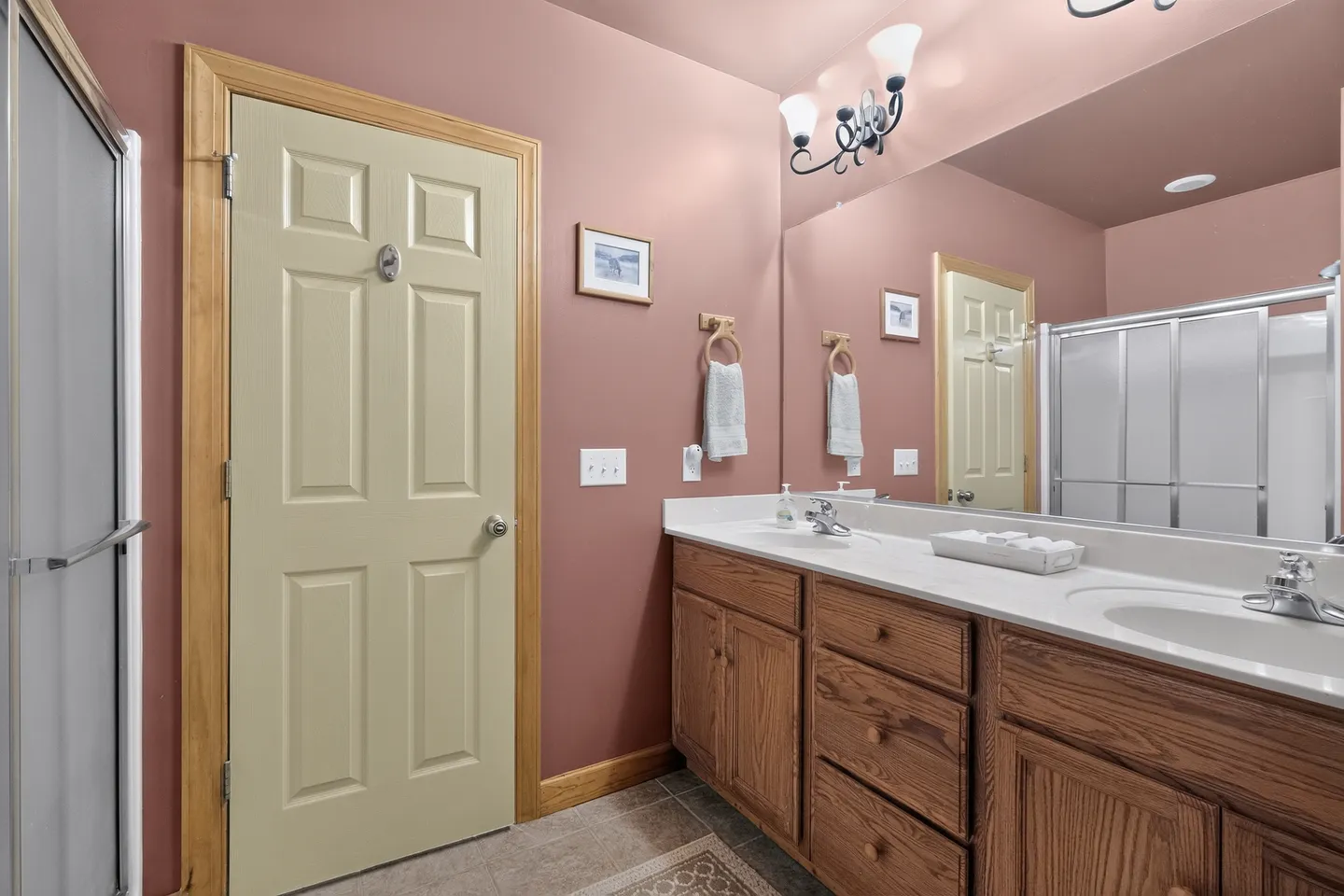 Bathroom with pink walls, wood double vanity, large mirror, and a frosted glass shower door.