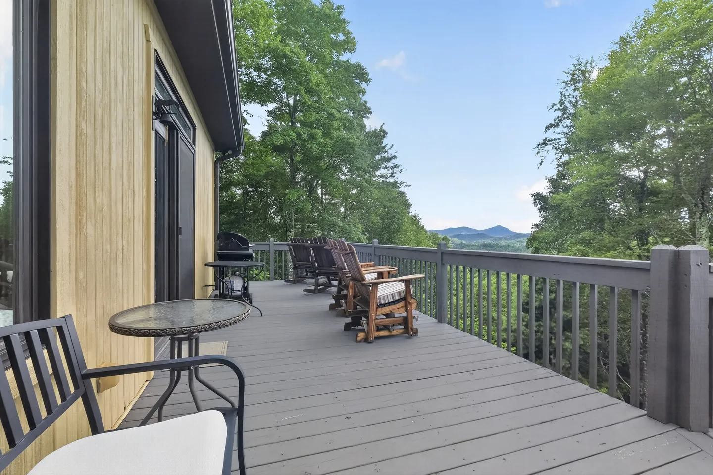 Elevated wooden deck with railing, outdoor seating, a small table, a grill, and views of distant mountains and trees.