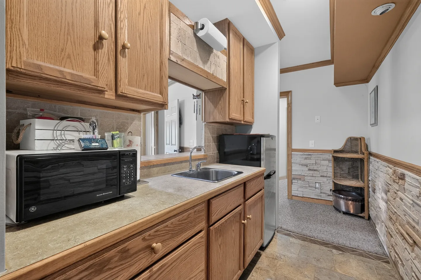 Kitchenette area with wood cabinets, countertop with a microwave, small sink, and mini fridge next to a hallway.