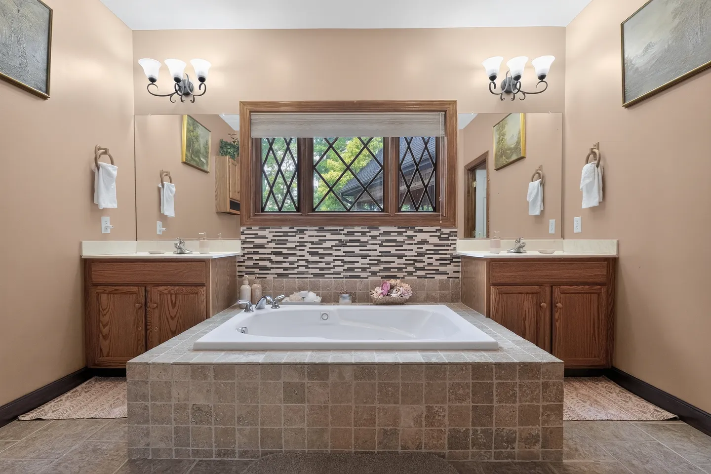 Bathroom with a large tiled jetted bathtub centered between dual wood vanities with mirrors and overhead lights.