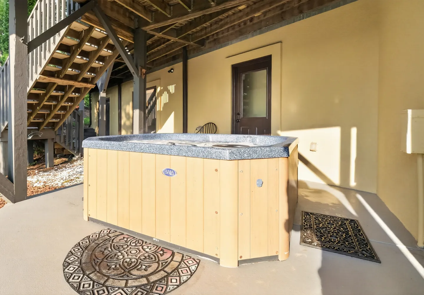 A covered outdoor patio featuring a hot tub beneath a wooden deck and staircase.
