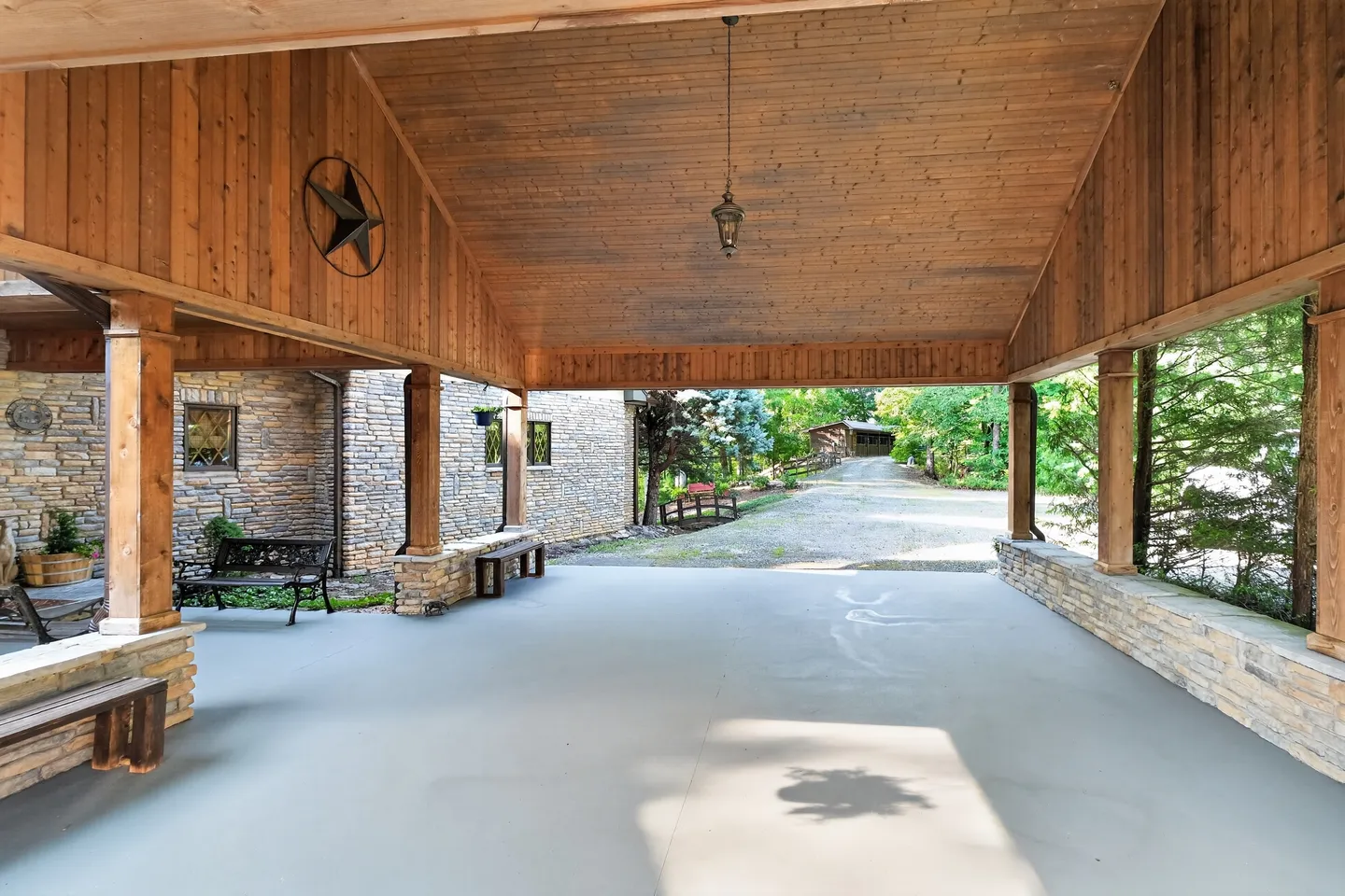 A covered outdoor carport or pavilion with wood-paneled walls and ceiling, stone accents, and benches overlooking a gravel driveway.