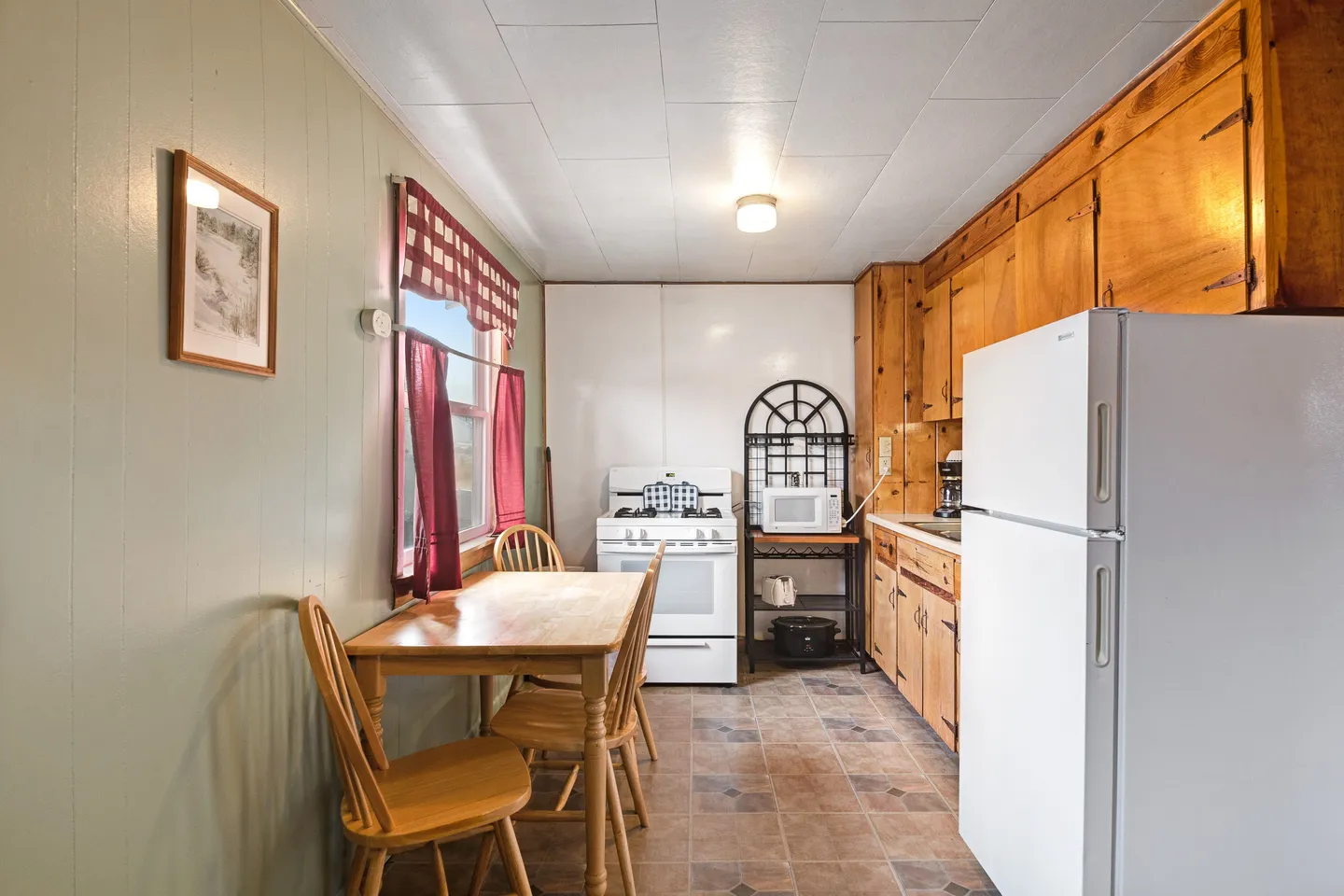 Kitchen with wooden cabinetry, a white refrigerator, gas stove, and a dining table with chairs under a window with red checkered curtains.