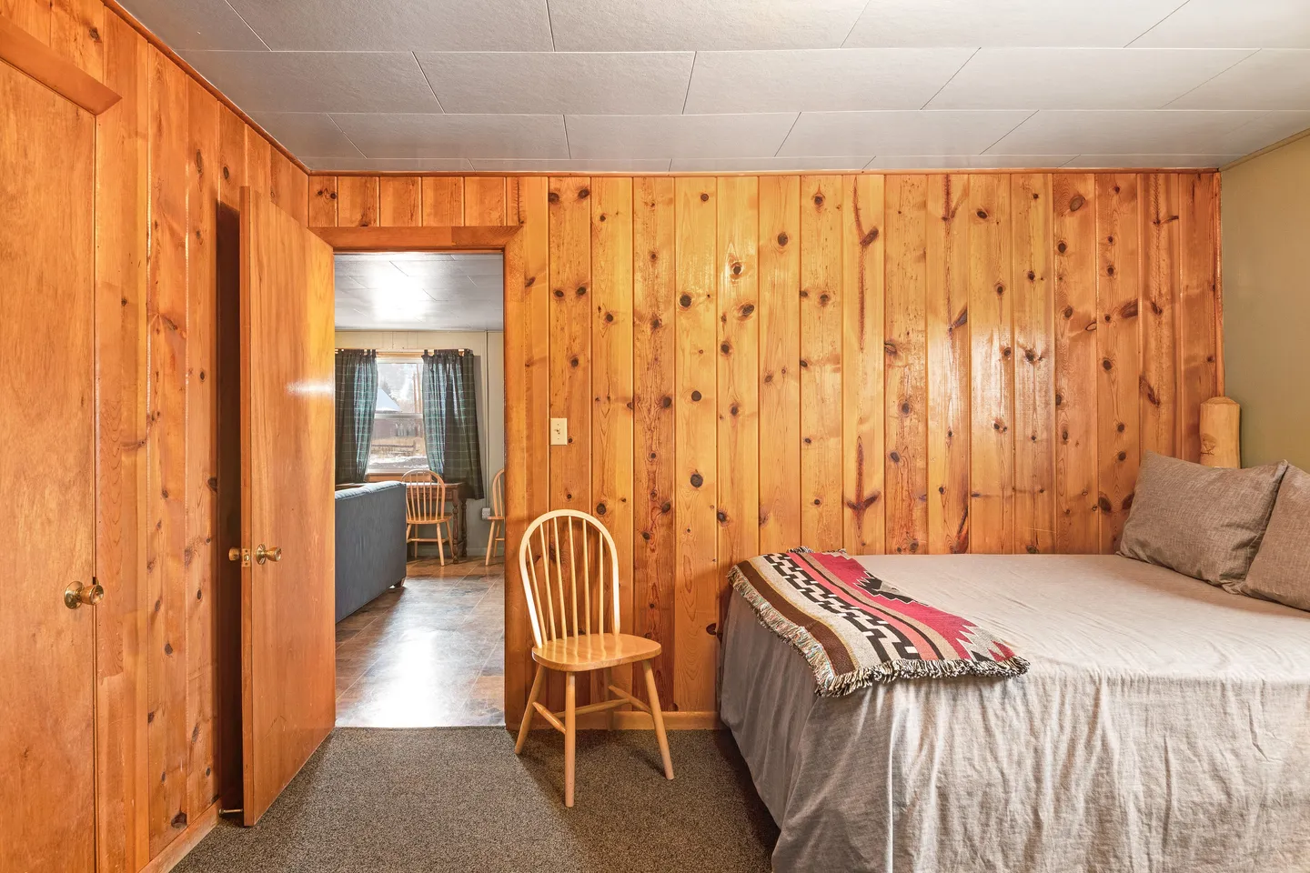 Bedroom with knotty pine wood-paneled walls, carpeted floor, a bed, a wooden chair, and an open doorway showing a living area.