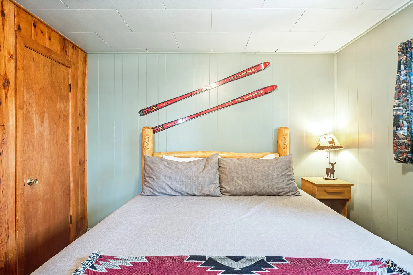 Bedroom with a log headboard bed, pair of red skis mounted on the wall, wood-paneled door, and small nightstand with a lamp.