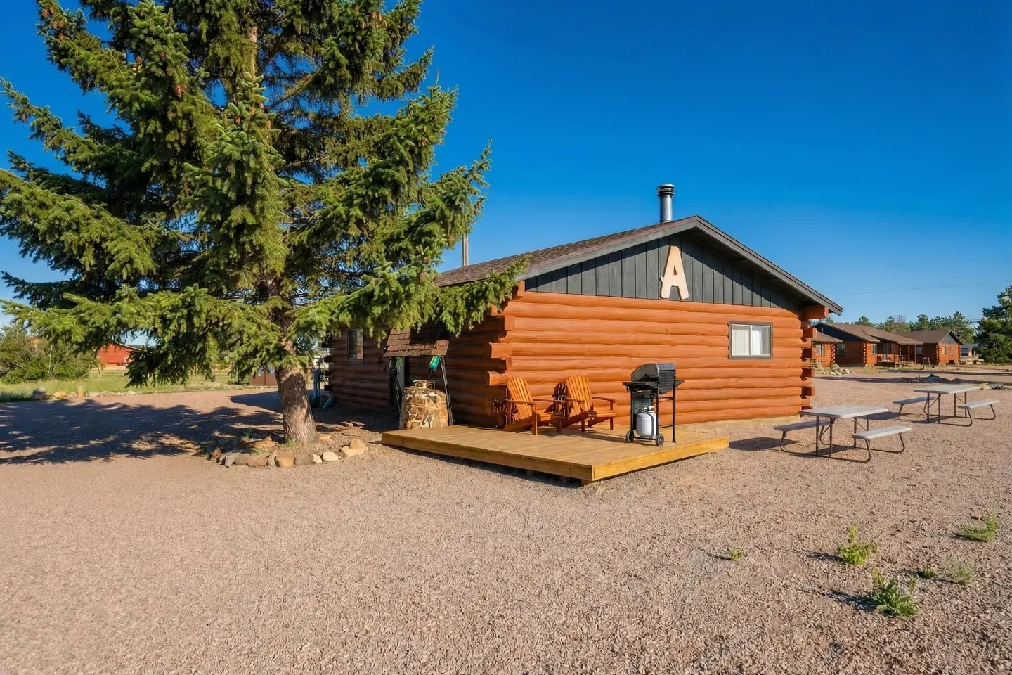 Exterior of a log-sided cabin labeled with the letter A, featuring a small wooden deck with chairs, a gas grill, and a picnic table on gravel grounds under a clear blue sky.
