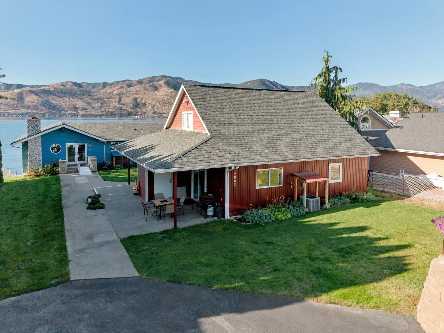 Elevated view of a red-sided house with a patio, lawn, and lake with mountains in the background under clear skies.