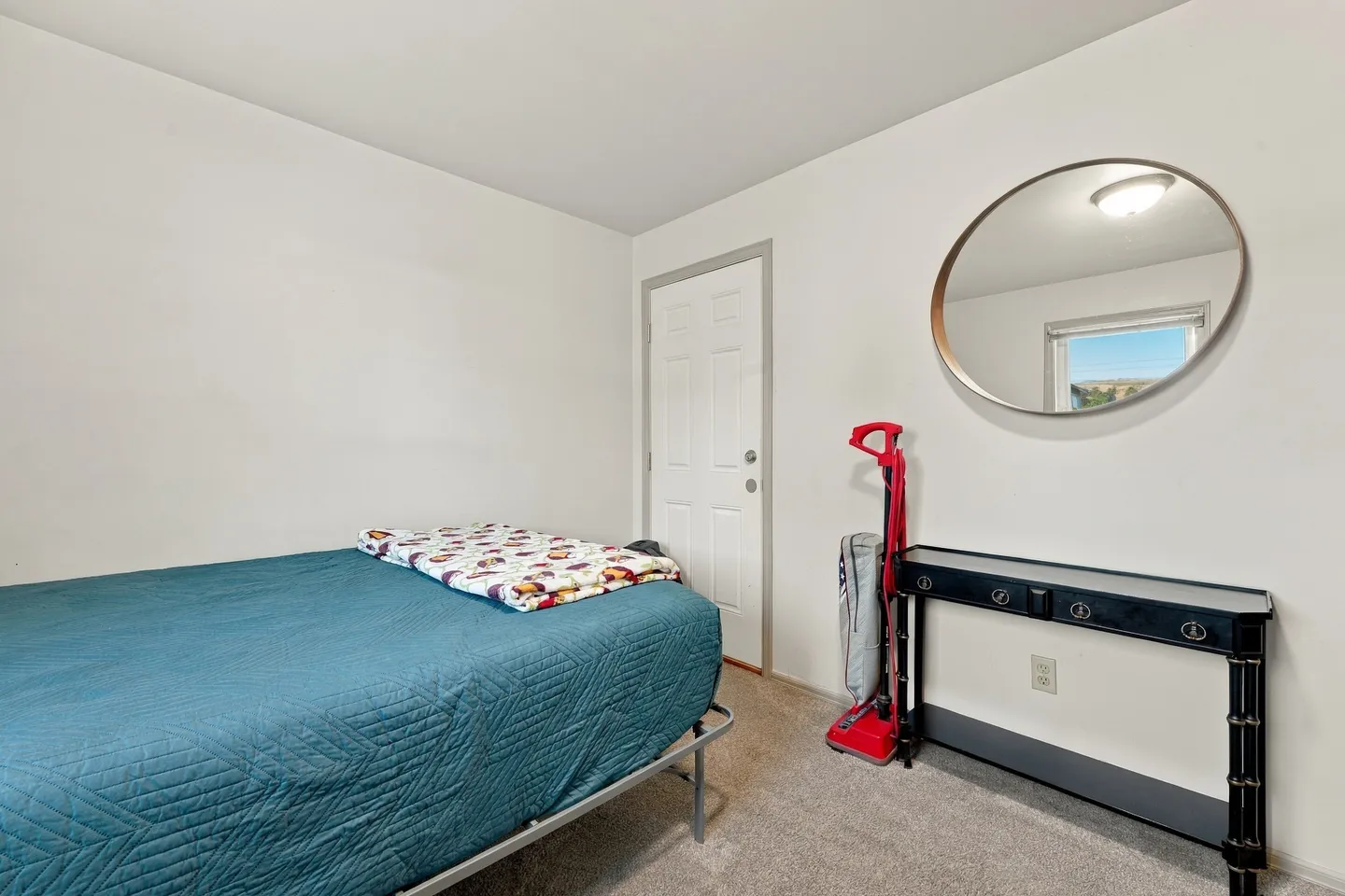 Bedroom with a bed made up with a teal quilt, a dark console table under a round mirror, and a door.