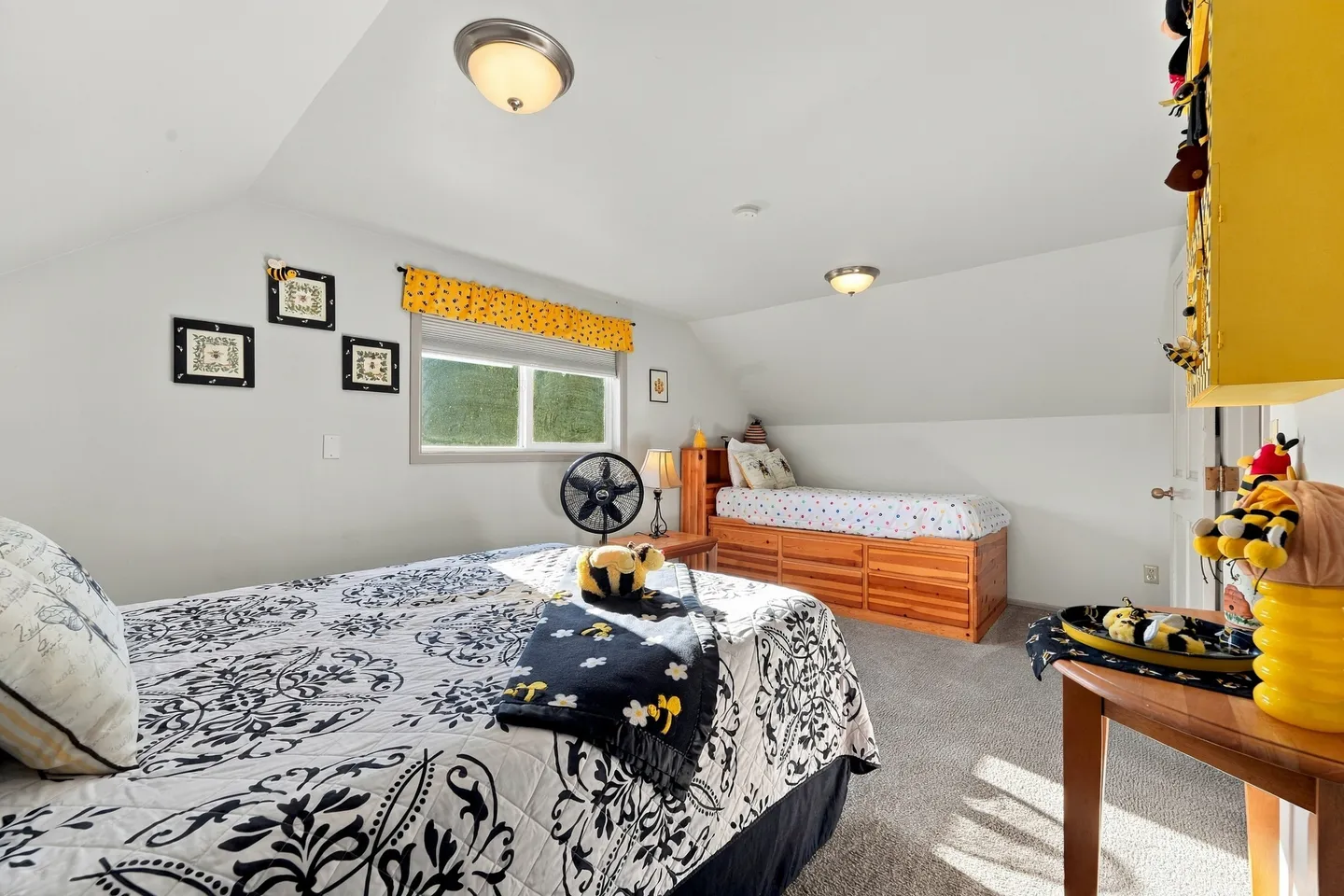 Bedroom with sloped ceiling, carpeted floor, a bed with black and white patterned quilt in foreground, and a single bed with drawers in the corner.