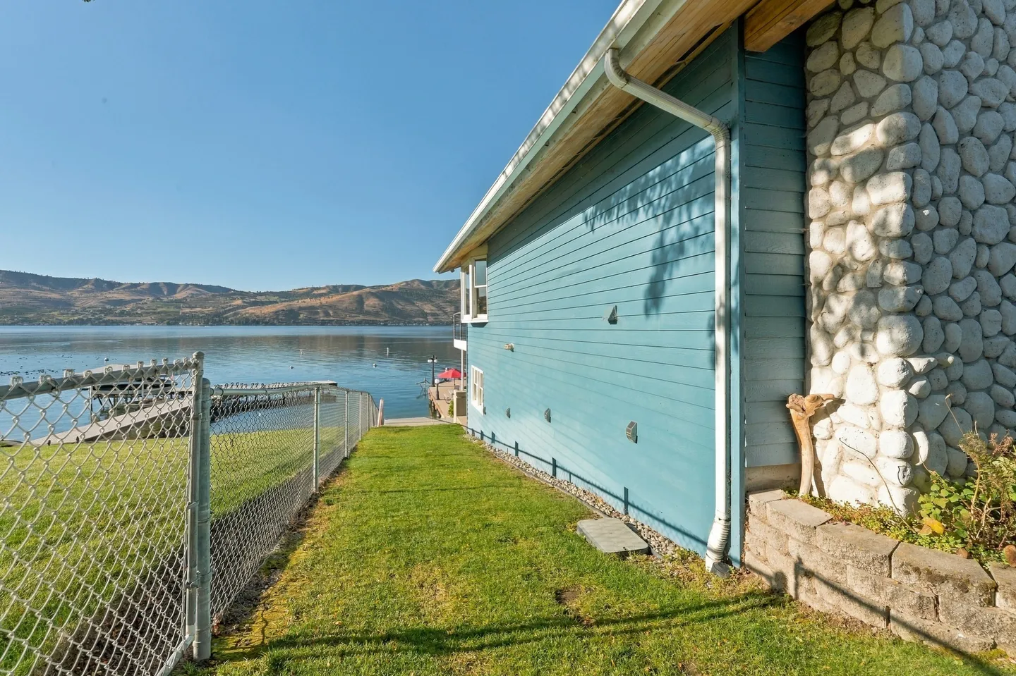 A grassy side yard next to a blue house with a stone chimney, leading down toward a chain-link fence, dock, and lake with distant hills.