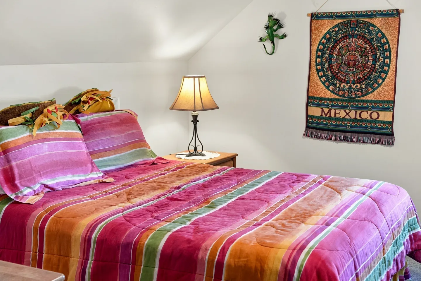 Bedroom featuring a bed with a colorful striped quilt and pillowcases, a nightstand with a lamp, and Mexican-themed wall decor.
