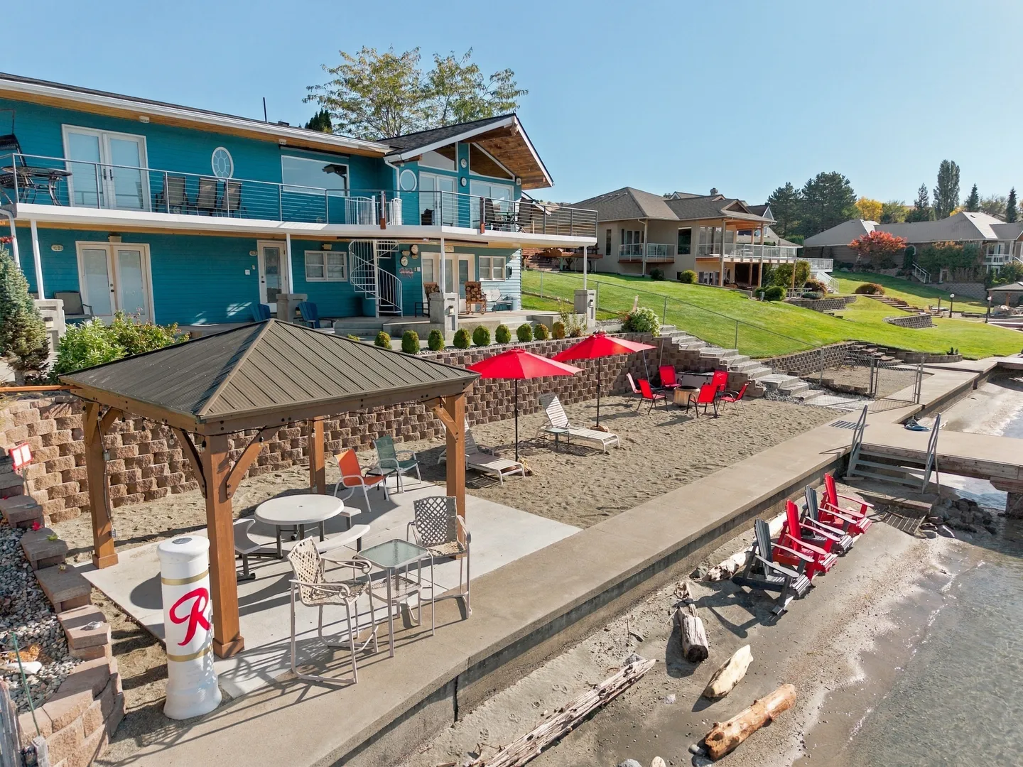 A high-angle view of a blue waterfront rental home with a multi-level outdoor seating area, gazebo, sand terrace, and shoreline.